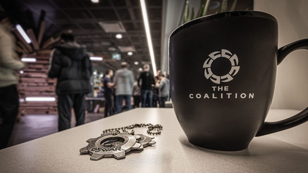 A mug with the coalition logo and metal cog tag necklace laying on a table with people blurry in the background.